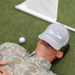 Person lying on grass with a golf ball and club, wearing a cap with 'High Creek' branding.