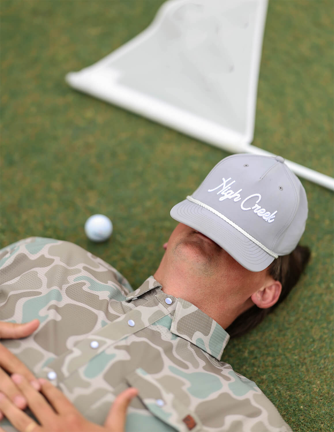 Person lying on grass with a golf ball and club, wearing a cap with 'High Creek' branding.